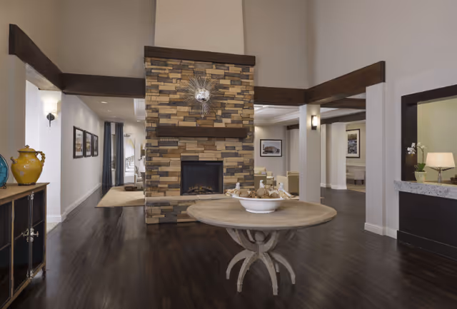 Interior view of a senior living facility lobby featuring a central stone fireplace with a decorative sunburst mirror above it. In front of the fireplace is a round wooden table with a bowl of decorative items. The room has dark wood flooring, white walls, and wooden beams. To the left, there is a cabinet with decorative pottery, and to the right, a reception desk with a lamp and a small plant. Seating areas and framed pictures are visible in the background.