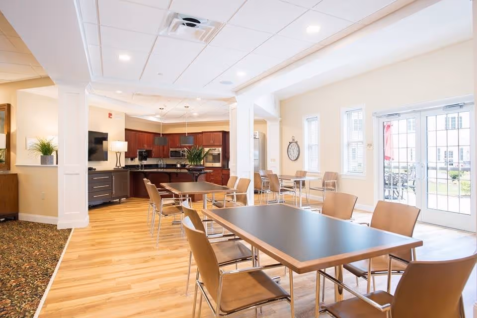 Bright communal dining and kitchen area with multiple tables and chairs, wood floors, and glass doors to the outside.
