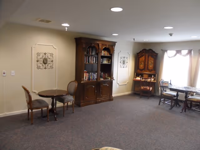 A cozy interior room with carpeted floor featuring a small round table with two chairs on the left, a large wooden bookshelf filled with books in the center, and a wooden cabinet with a chair and a table with chairs near a window with sheer curtains on the right. The walls are light-colored with decorative wall panels and ceiling lights.