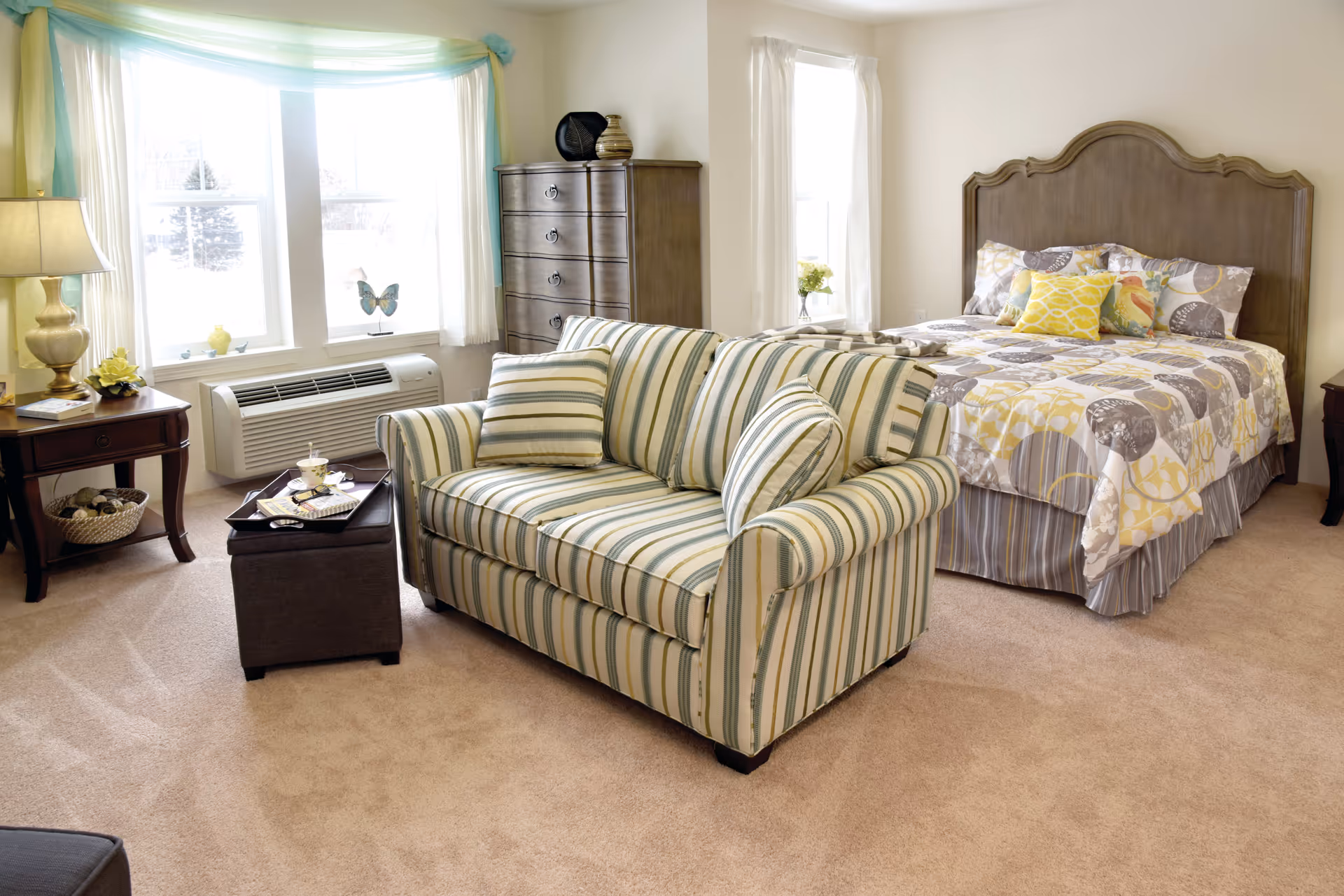 A bright and cozy bedroom featuring a bed with patterned bedding and multiple pillows, a striped loveseat with matching cushions, a wooden dresser, a side table with a lamp and decorative items, and large windows with sheer curtains allowing natural light to fill the room.