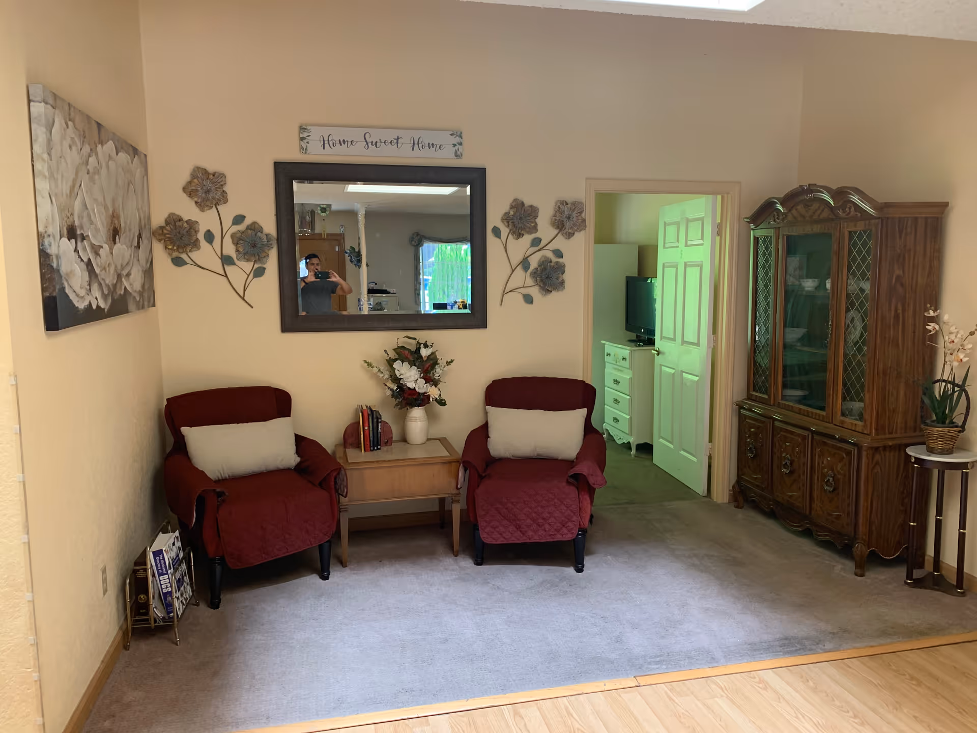A cozy living room area with two red armchairs covered with quilted throws and beige cushions. Between the chairs is a small wooden side table with a vase of flowers and some books. Above the table is a large mirror with a 'Home Sweet Home' sign above it and decorative metal flower wall art on either side. To the right is a wooden china cabinet with glass doors displaying dishes, and a small round table with a potted plant. The room has beige walls and carpeted flooring, with a doorway leading to another room where a TV and dresser are visible.