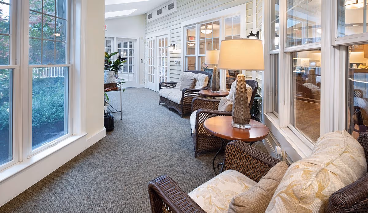 A bright and cozy indoor seating area with wicker armchairs featuring floral cushions, round wooden side tables with decorative lamps, large windows letting in natural light, and a view of greenery outside.