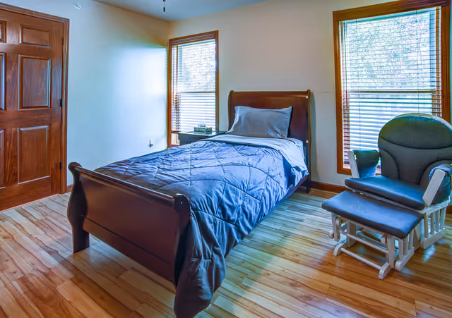 A simple bedroom with a wooden single bed covered with a blue quilt and a gray pillow. There are two windows with wooden blinds letting in natural light. Next to the bed is a small nightstand with a few books on it. A cushioned rocking chair with an ottoman is placed near one of the windows. The room has wooden flooring and a wooden door.