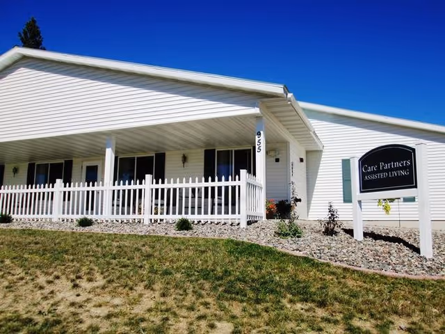 White single-story assisted living building with a covered porch, white picket fence, front lawn, and a sign reading 'Care Partners Assisted Living'.