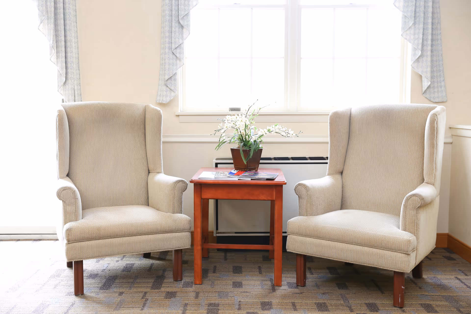 Two beige upholstered armchairs with wooden legs are positioned on either side of a small wooden table with a potted plant and some magazines on top. Behind the chairs is a large window with light-colored curtains, and the floor is carpeted with a patterned design.