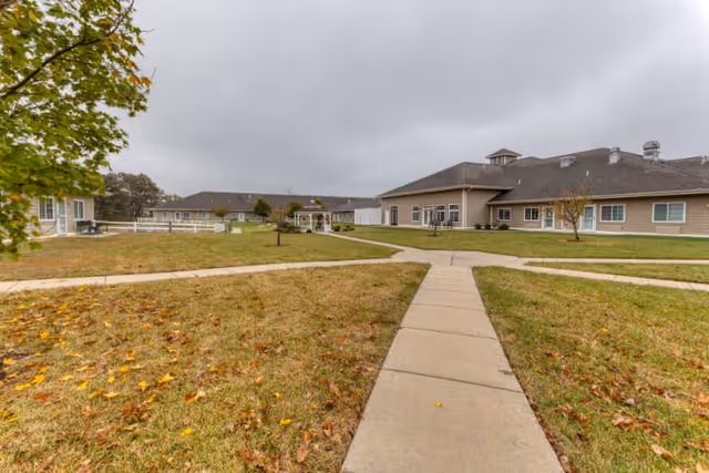 Outdoor view of Cedarhurst Senior Living of Waterloo showing a paved walkway intersecting grassy areas with a few small trees, surrounded by single-story beige buildings under a cloudy sky.