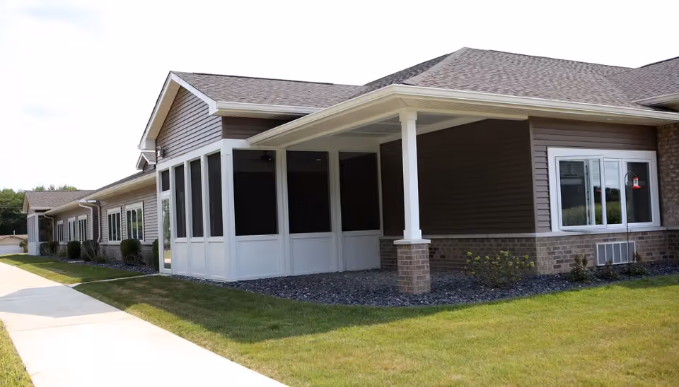 Exterior view of a single-story assisted living facility building with brown siding and brick accents. The building features a covered porch area with white columns and screened windows. There is a concrete walkway and well-maintained grass surrounding the building.