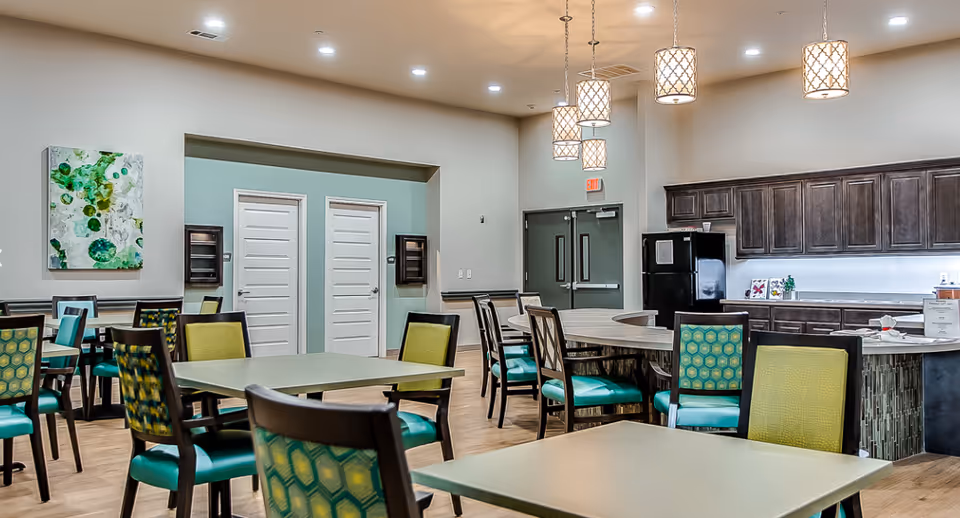A spacious dining room in a senior living facility featuring multiple tables and chairs with green and yellow patterned upholstery. The room has a modern design with pendant lights hanging from the ceiling, a kitchen area with dark wooden cabinets, a black refrigerator, and a countertop. There is a green and white abstract painting on the wall and two white doors in the background.