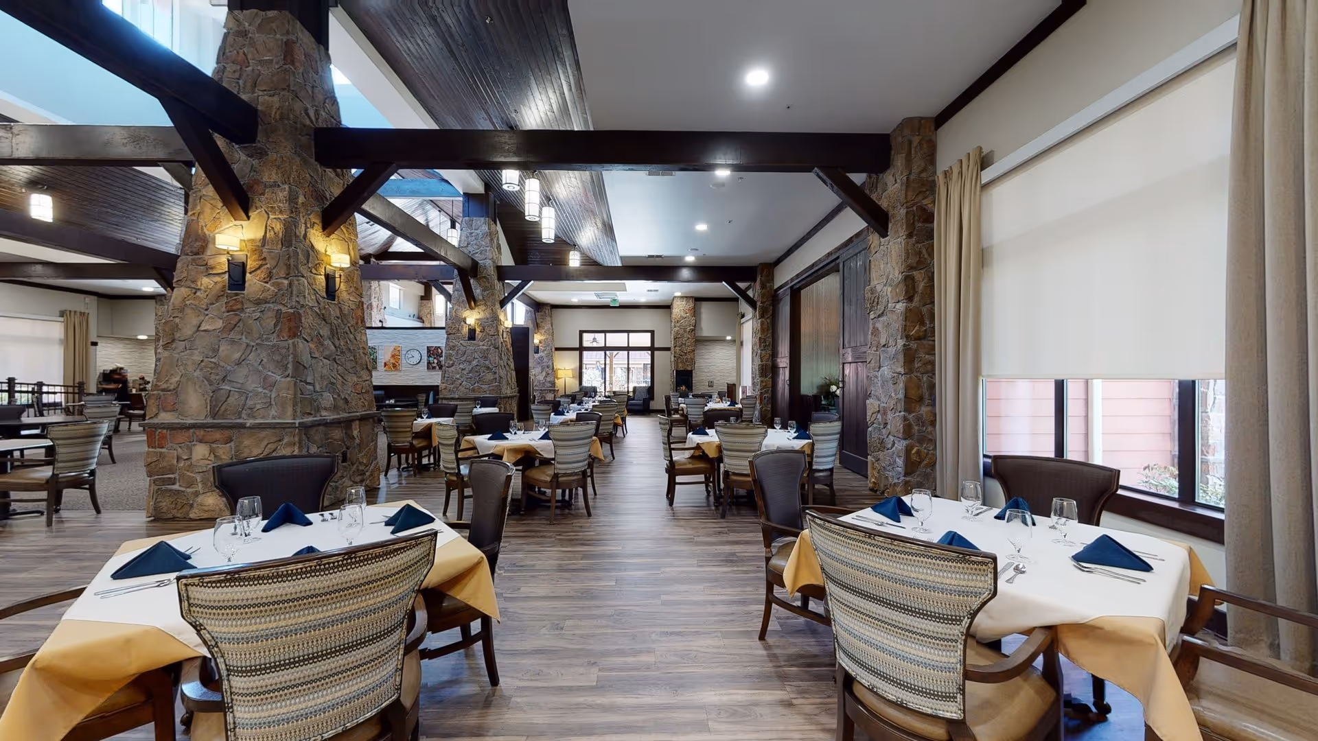 Spacious dining room with multiple tables set with white tablecloths, yellow overlays, blue napkins, glassware, and silverware. The room features stone pillars, wooden beams on the ceiling, large windows with curtains, and wood flooring.