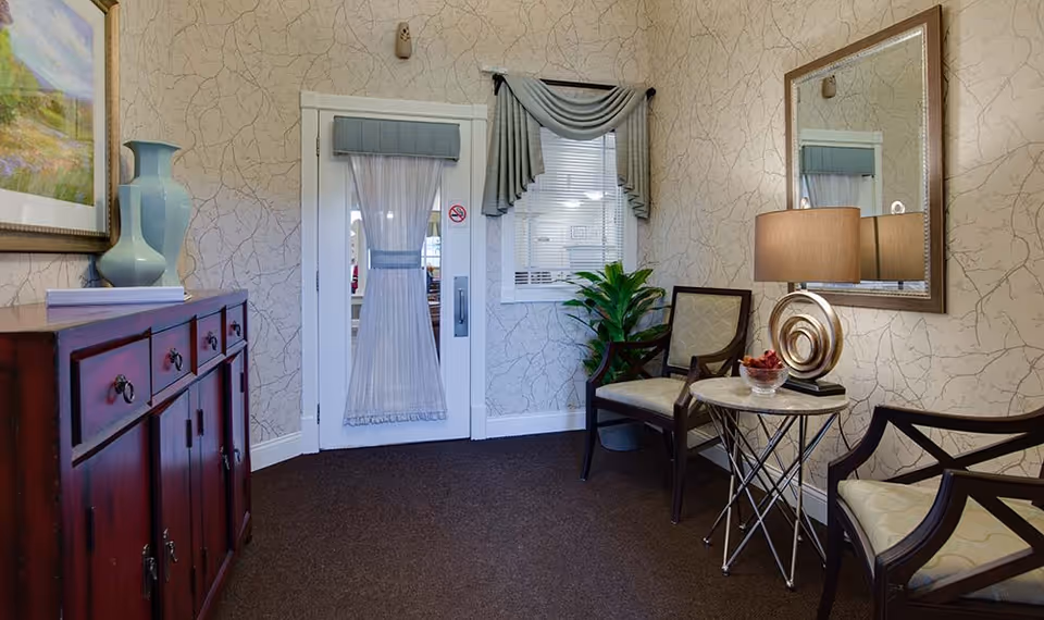 A cozy interior sitting area with two cushioned wooden chairs around a small round table holding a decorative lamp and a bowl of potpourri. A large mirror hangs on the wall above the table. To the left, there is a wooden cabinet with a blue vase and a framed painting above it. A door with a sheer curtain and a window with a valance are visible in the background, along with a potted plant near the chairs.