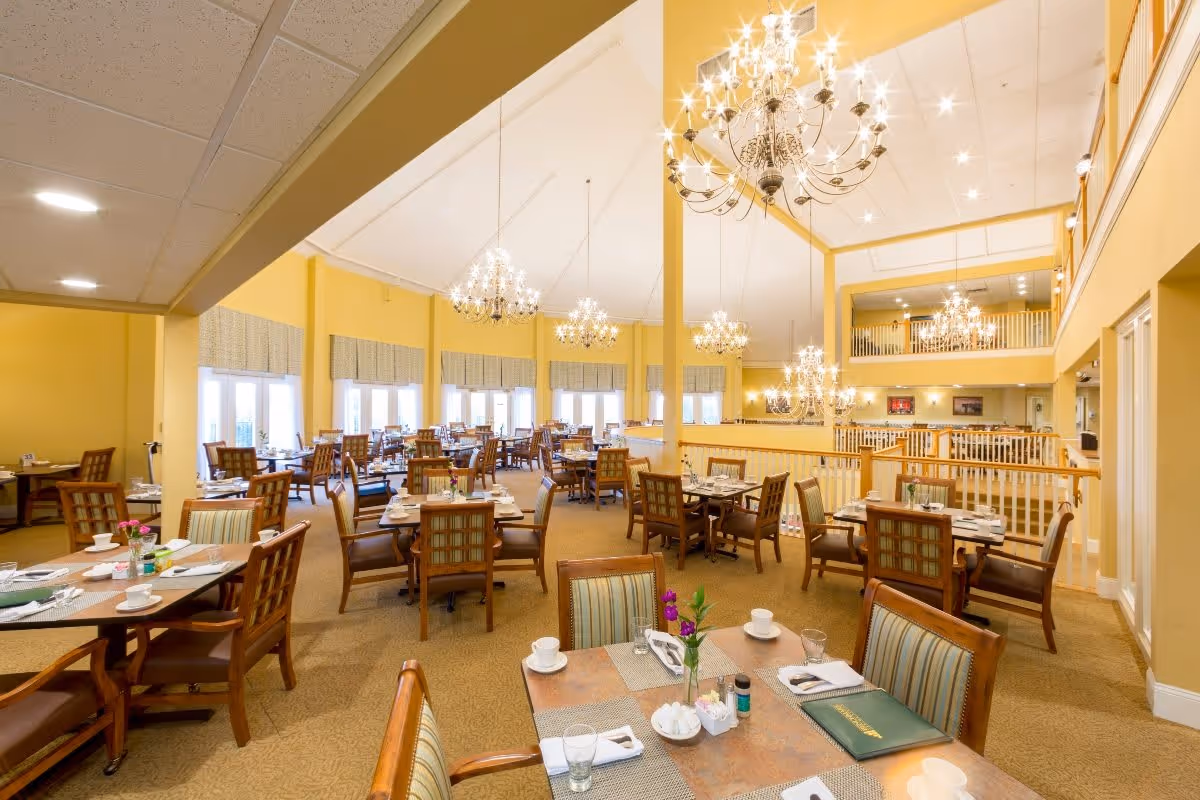 A spacious, well-lit dining room with multiple wooden tables and chairs arranged neatly. Each table is set with placemats, napkins, cups, and small flower vases. The room features large windows with valances, high ceilings with elegant chandeliers, and a warm yellow color scheme.