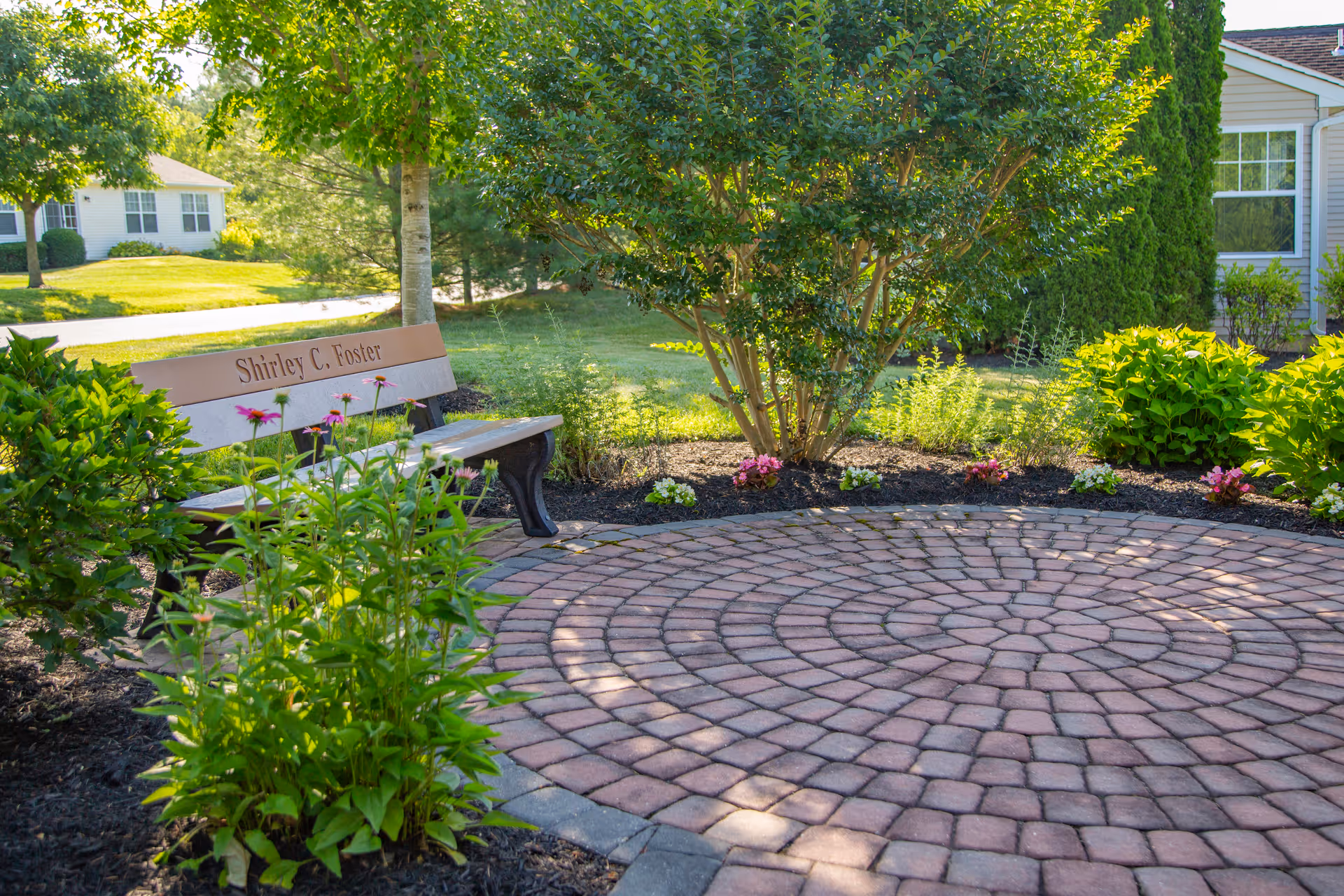 Circular brick patio with a bench engraved "Shirley C. Foster" surrounded by shrubs and flowering plants in a sunny garden.