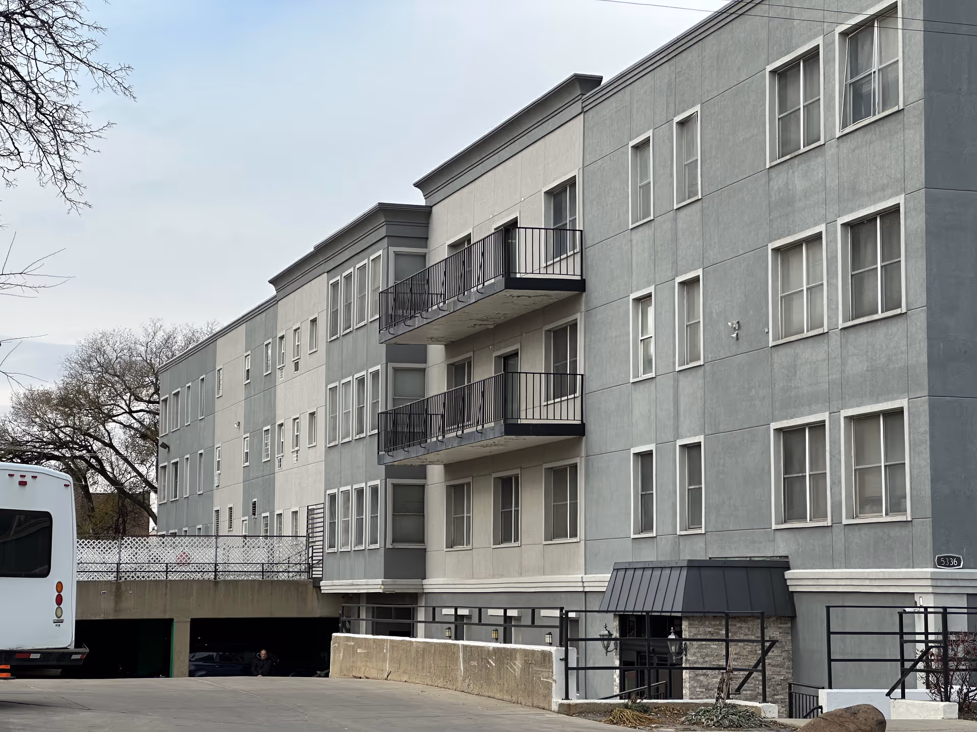 Exterior view of a multi-story gray building with multiple windows and two balconies. There is a driveway leading to a parking garage entrance, and a white vehicle is partially visible on the left side. Leafless trees are visible in the background under a cloudy sky.