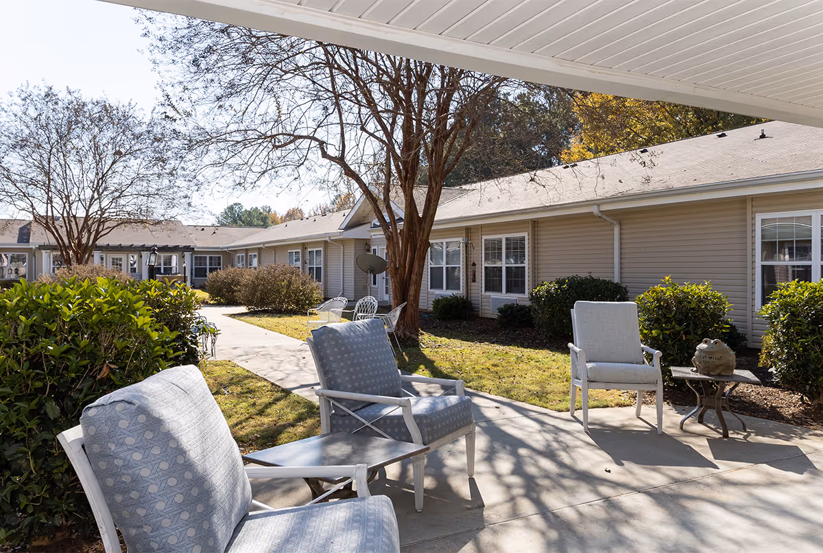 Outdoor patio area at Hammond Square with cushioned chairs and a small table under a covered walkway. The patio overlooks a garden with bushes, trees, and a beige single-story building with multiple windows in the background.