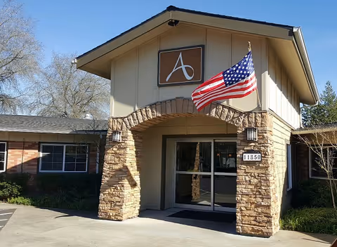 Entrance of a building with stone pillars and an archway, featuring a sign with a stylized letter 'A' above the door and an American flag mounted on the right pillar. The building has beige siding and a dark roof, with windows visible on the left side and some greenery around the base.