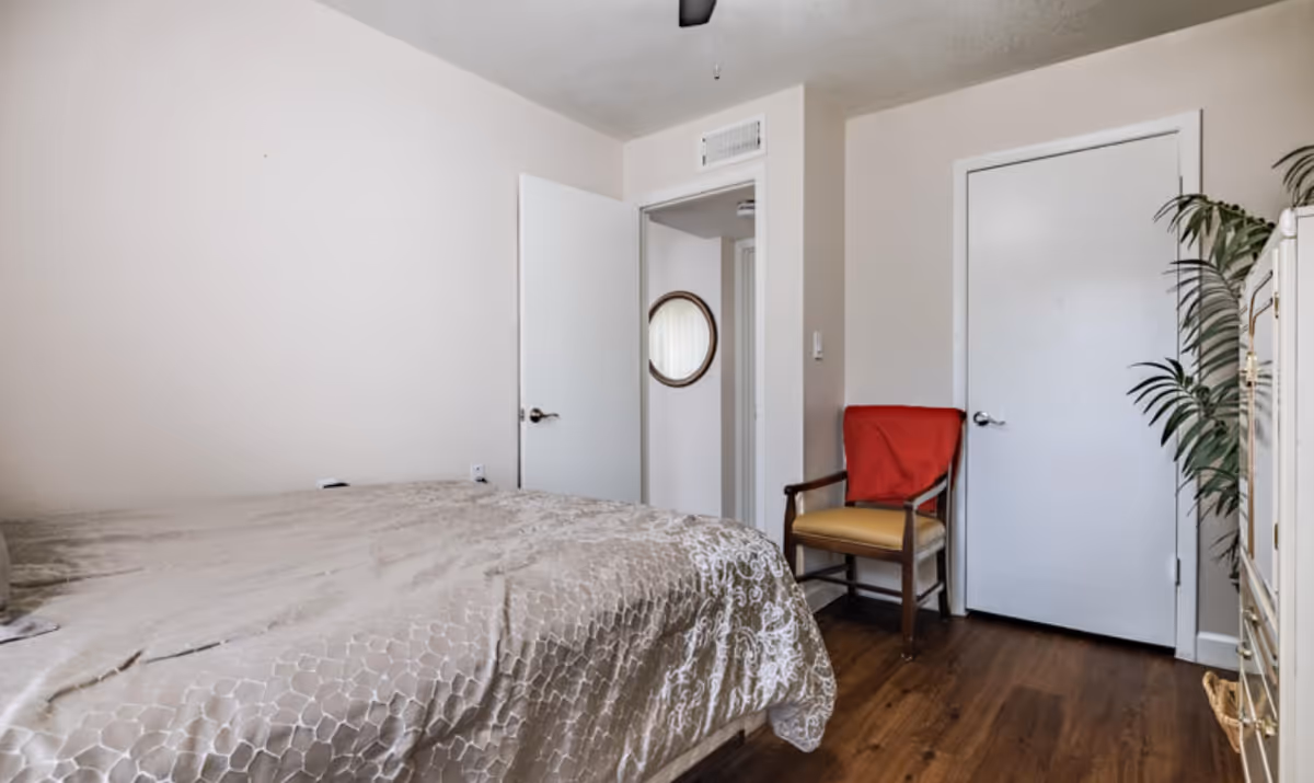 Simple bedroom with a bed covered in a patterned comforter, a wooden chair with a red throw, two doors, and hardwood flooring.