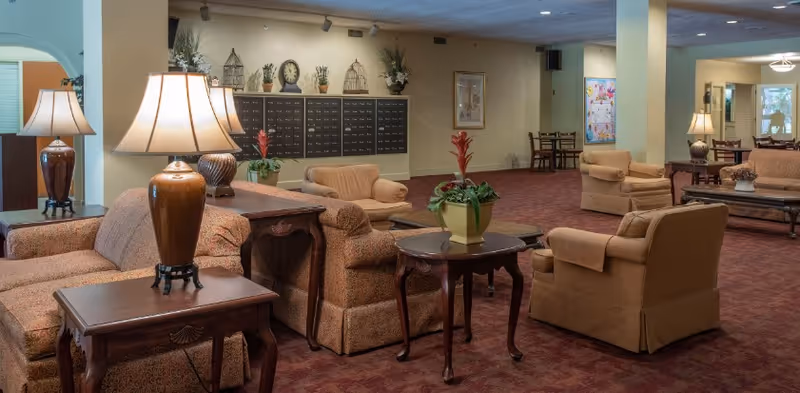 A spacious living room area with multiple upholstered armchairs and sofas arranged around wooden side tables with lamps. There are decorative plants on the tables and a wall with mailboxes and clocks in the background. The room has a carpeted floor and soft lighting.