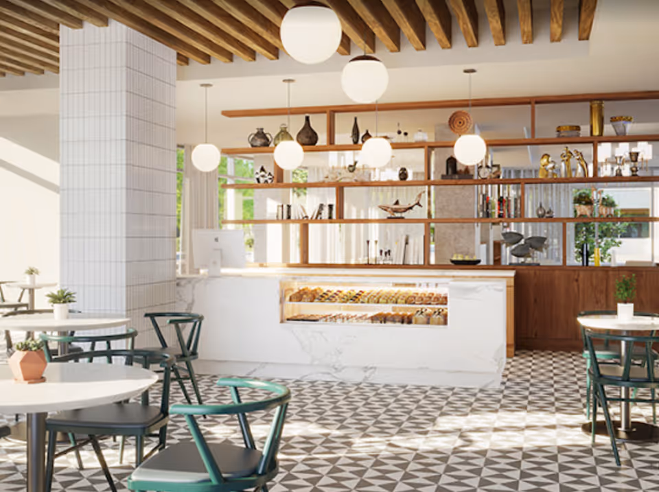 Bright and modern dining area with round white tables and green chairs, featuring a white marble counter displaying pastries. The space has geometric patterned floor tiles, wooden ceiling beams, hanging globe pendant lights, and wooden shelves with decorative items.