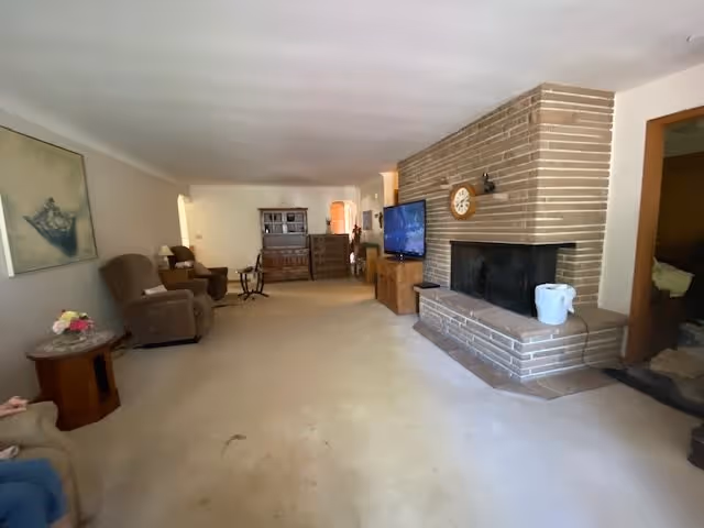 A spacious living room with beige carpet and walls. On the right side, there is a large brick fireplace with a clock mounted above it and a white bucket placed on the hearth. Next to the fireplace is a wooden TV stand with a flat-screen television. On the left side, there are two brown recliner chairs and a small wooden side table with a flower arrangement. In the background, there is a wooden cabinet and other furniture pieces along the far wall. The room has a cozy and traditional feel.