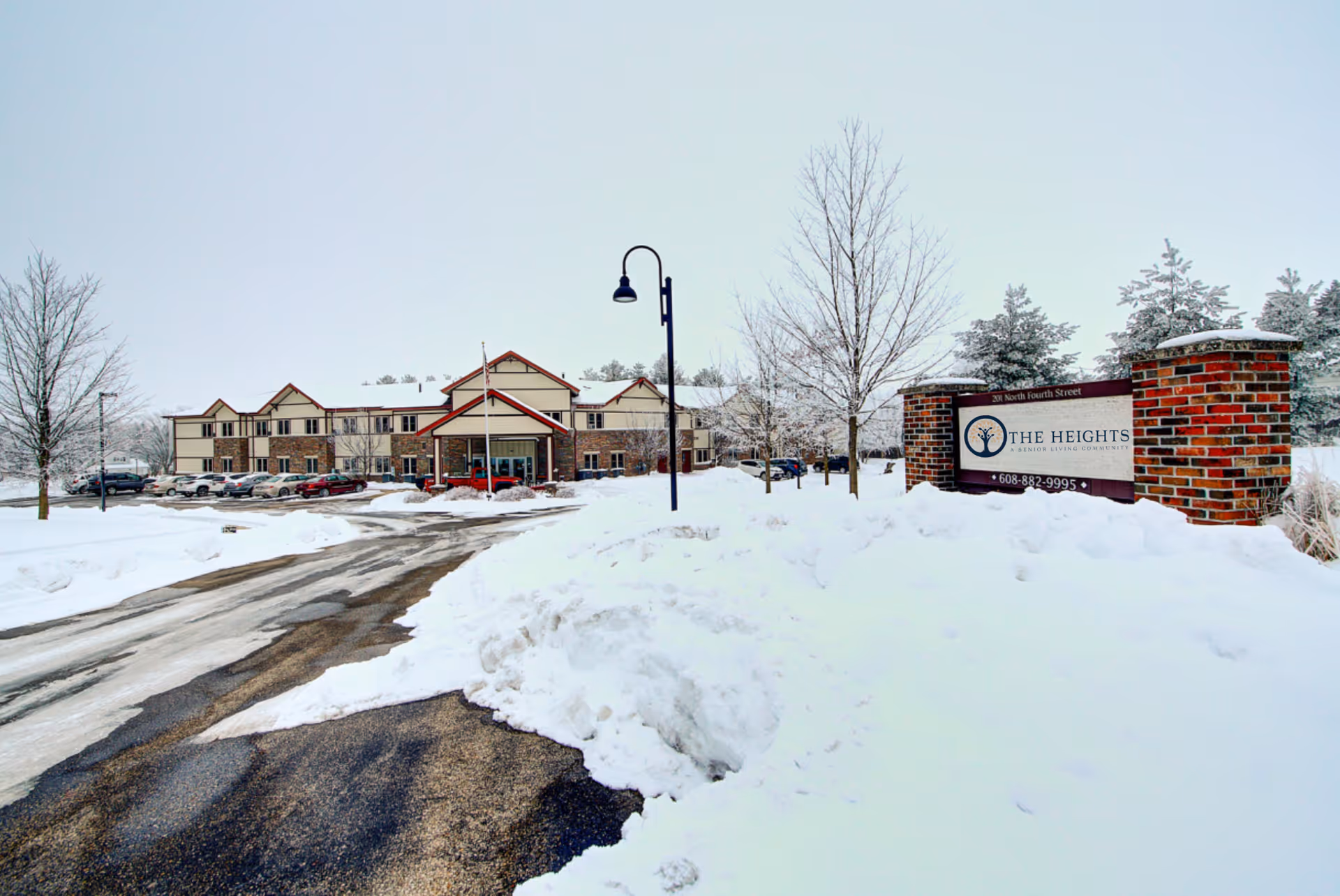 Snow-covered driveway leading to a two-story senior living community building with a red roof and beige exterior. There are several parked cars along the driveway, leafless trees, and a brick sign on the right side that reads 'The Heights, A Senior Living Community'. The ground and surrounding area are covered in snow.