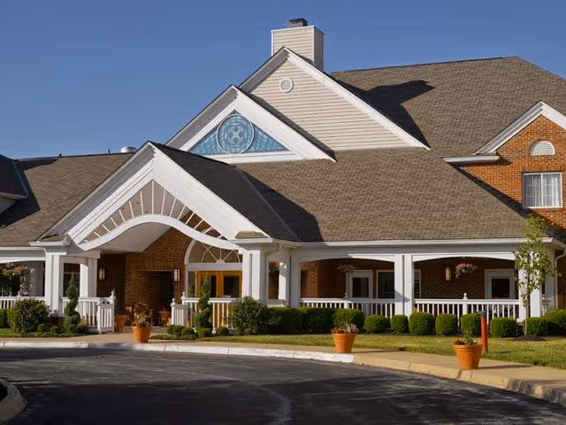 Front entrance of a brick residential building with a white-pillared covered portico, potted plants, and manicured landscaping under a clear blue sky.