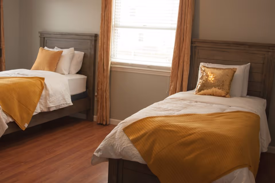 A bedroom with two single beds featuring wooden headboards, white bedding, and mustard yellow blankets and pillows. A window with mustard yellow curtains is centered between the beds, and the room has wooden flooring.