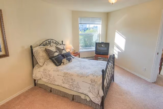 A bedroom with a metal frame bed covered with a white and beige floral bedspread and several pillows. There is a small wooden nightstand with a lamp next to the bed. A window with blinds lets in natural light, and a small television sits on a wooden stand in the corner. The room has beige walls and carpeted floor.