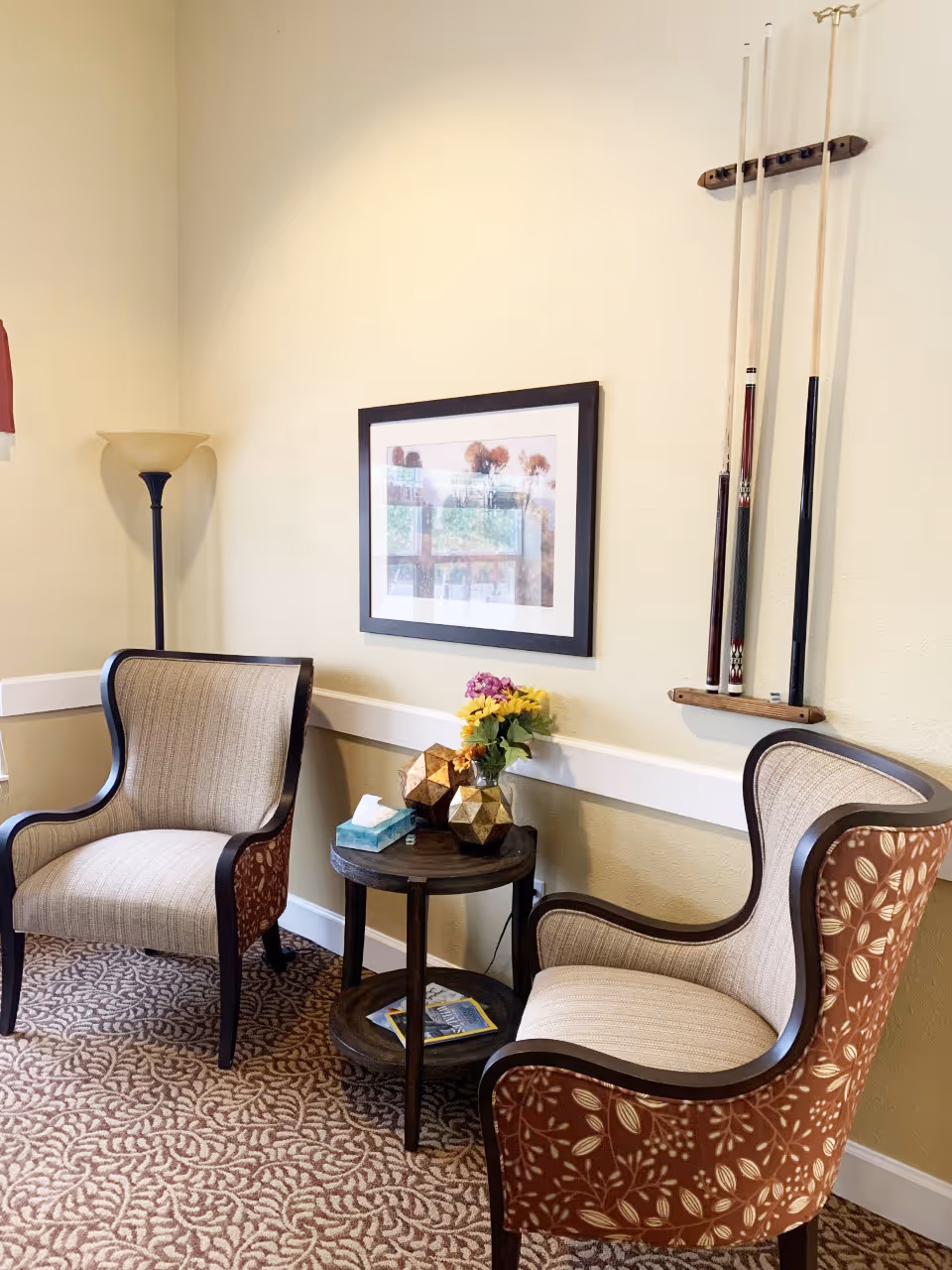 A cozy corner in a senior living facility with two upholstered armchairs featuring a beige and brown floral pattern. Between the chairs is a small round wooden table holding a box of tissues, decorative geometric objects, and a vase with yellow and purple flowers. On the wall above the table is a framed picture, and to the right, a rack holding three pool cues is mounted. A tall floor lamp stands in the corner, and the floor is covered with a patterned carpet.
