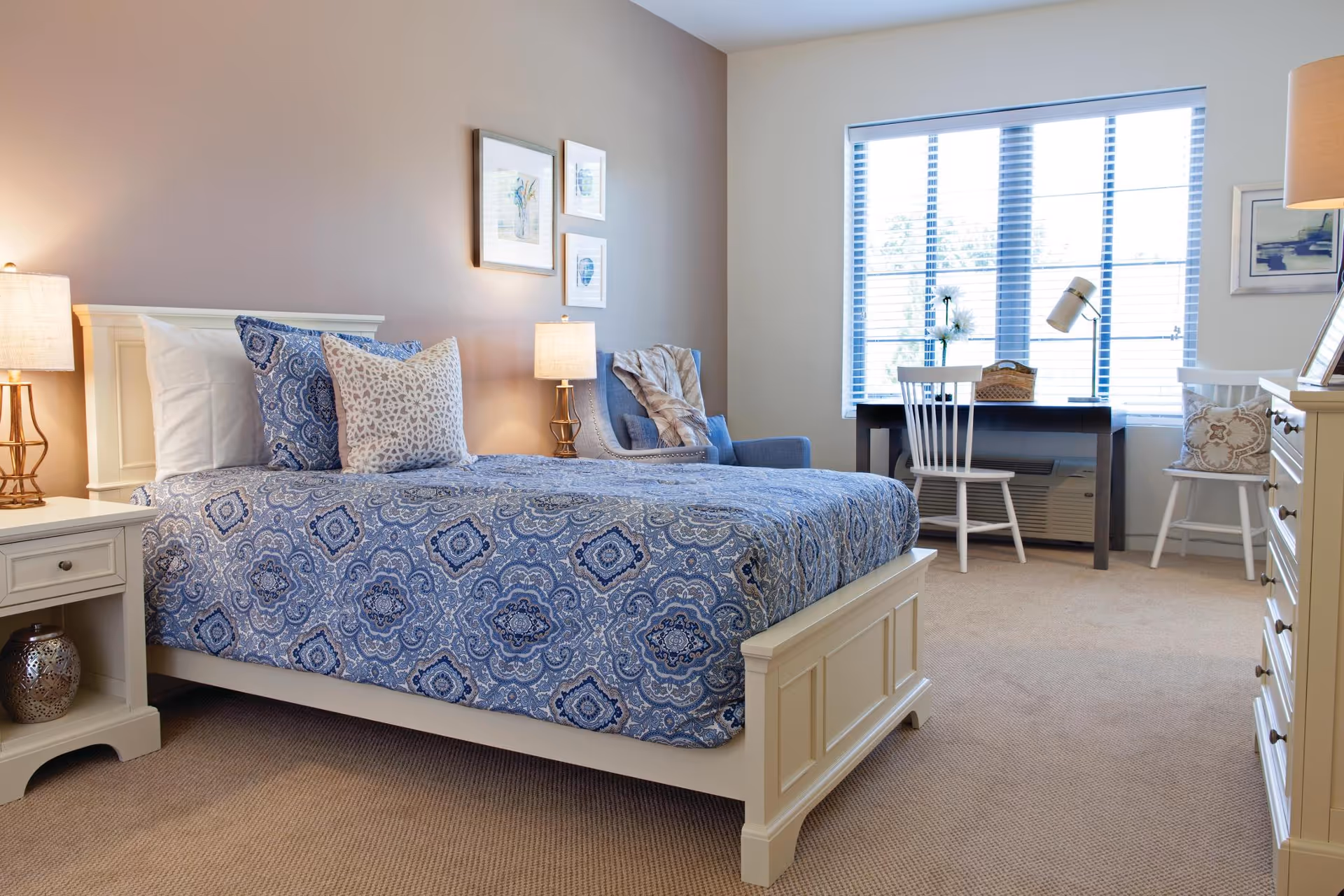 A bright and cozy bedroom with a white wooden bed frame and blue patterned bedding. The room features two bedside tables with lamps, a comfortable armchair with a throw blanket, a desk with a chair near a large window with blinds, and a white dresser. The walls are decorated with framed artwork.