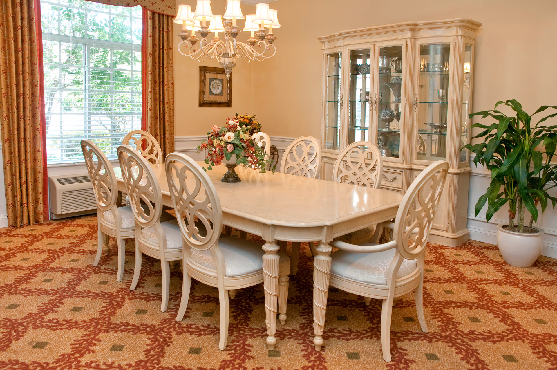 A formal dining room with a large rectangular table surrounded by eight ornate white chairs. A floral centerpiece sits on the table. The room features a patterned carpet, a large window with floral curtains, a chandelier overhead, a glass-fronted china cabinet, and a potted plant in the corner.