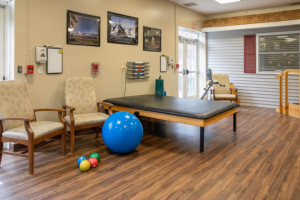 A therapy or rehabilitation room with wooden flooring, featuring a padded treatment table, three cushioned chairs with wooden arms, a large blue exercise ball, and four smaller colorful balls on the floor. The walls have motivational posters, a hand sanitizer dispenser, and other medical equipment. There is a door with an exit sign and a window with red shutters on the right side.