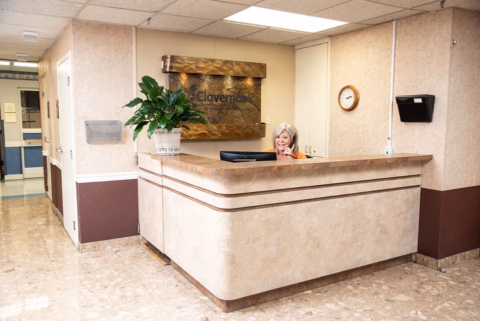 Reception area of Clovernook Health Care LLC with a smiling receptionist sitting behind a beige marble counter, talking on the phone. There is a potted plant on the counter, a clock on the wall, and a decorative sign with the facility's name behind the receptionist.