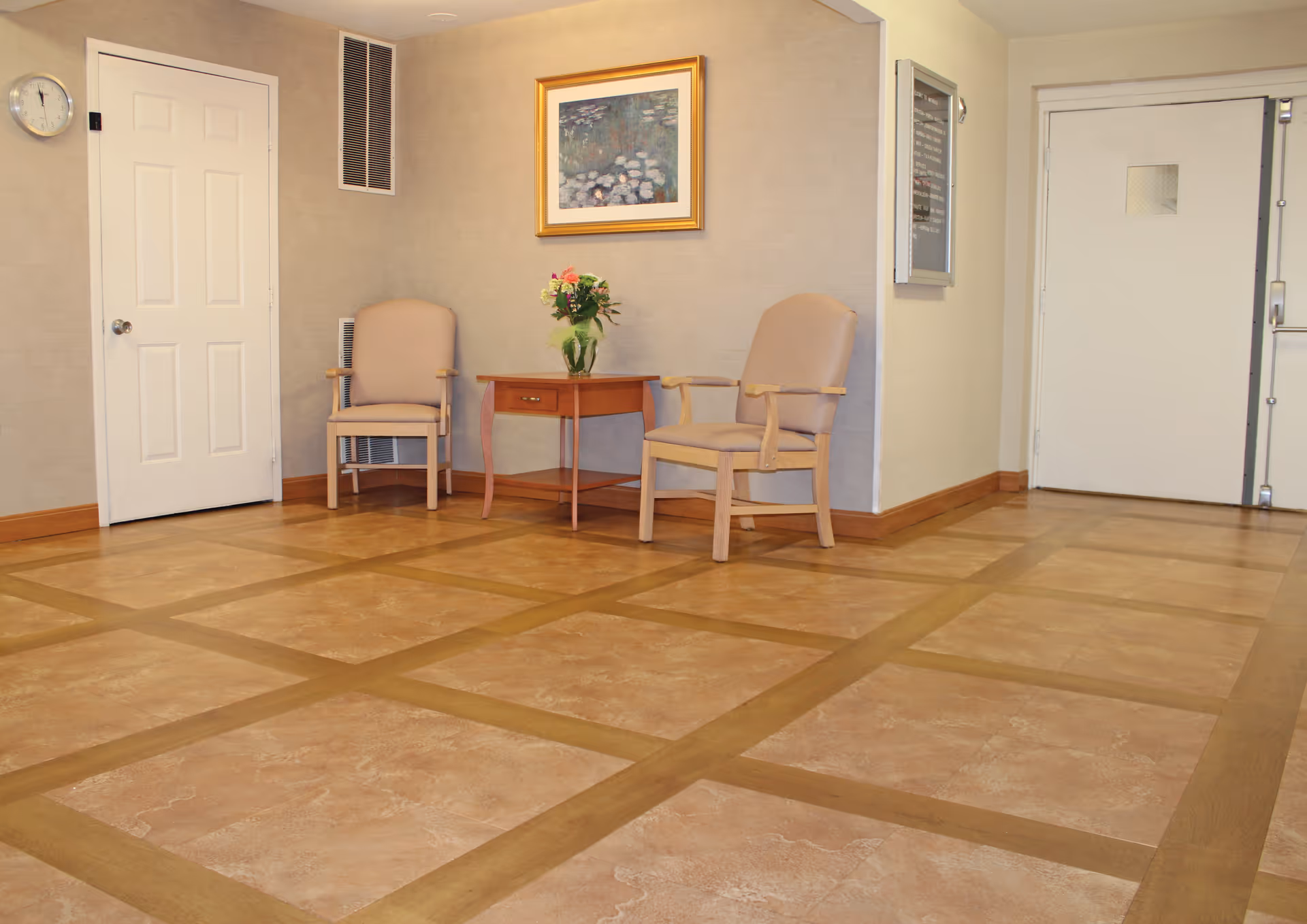 A waiting area with two beige armchairs and a wooden side table with a vase of flowers. The walls are light-colored with a framed painting hanging above the chairs. There is a white door on the left and another white door on the right side of the room. The floor has a patterned tile design.