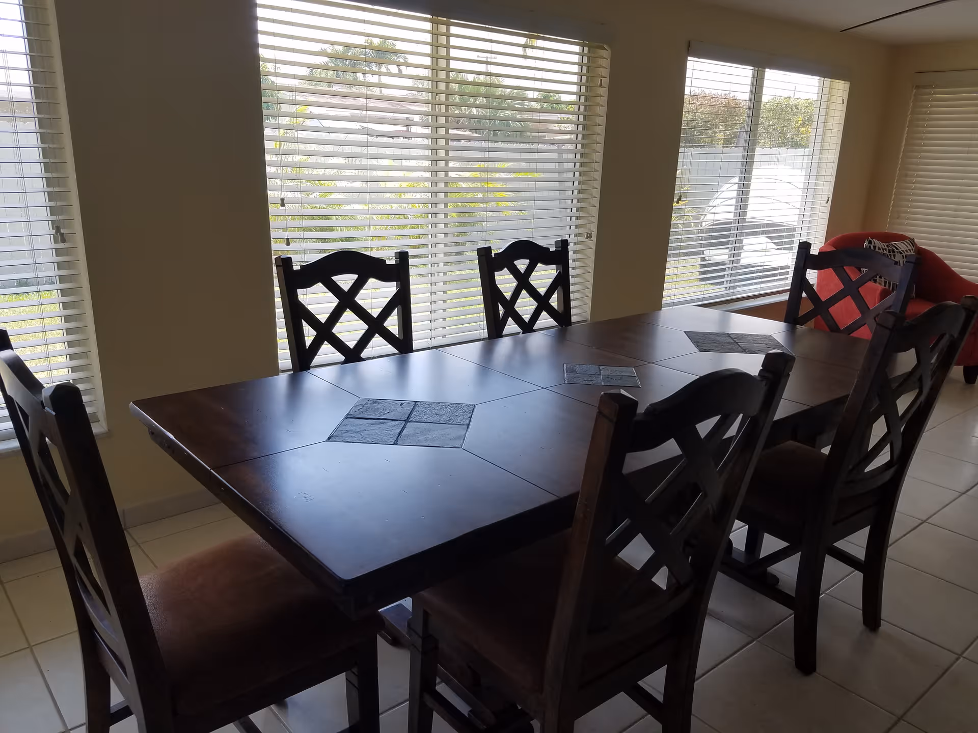 A dining area with a dark wooden rectangular table surrounded by six matching chairs. The table has decorative tile inlays. Large windows with white blinds allow natural light to fill the room. A red armchair is visible in the corner near the windows.