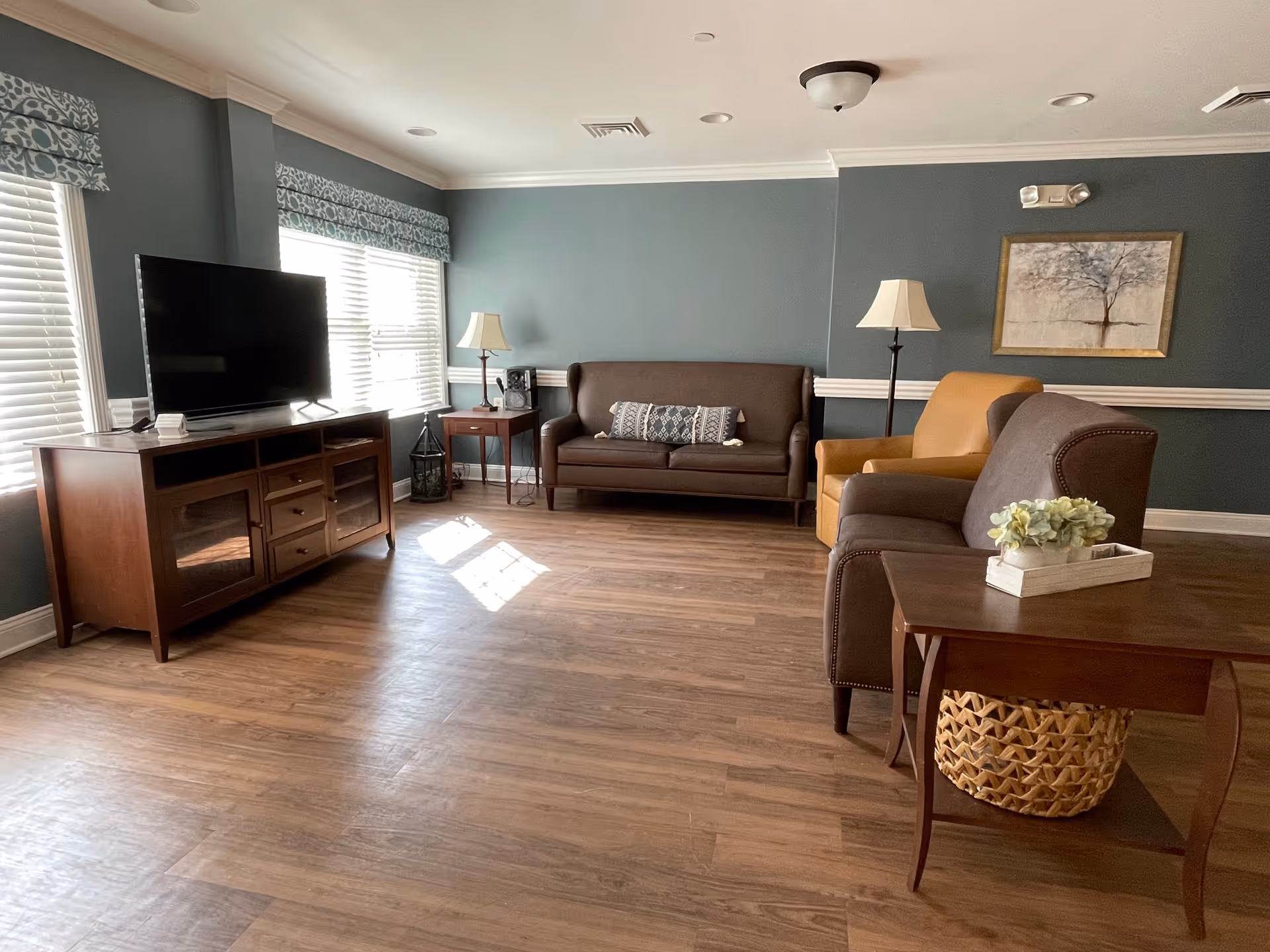 A cozy living room with wooden flooring, a brown sofa with a decorative pillow, a yellow armchair, and a brown armchair. There are two lamps, one on a side table and one floor lamp. A wooden TV stand holds a flat-screen TV. The walls are painted blue with white trim, and there is a framed picture of a tree on the wall. Windows with white blinds and patterned valances let in natural light.