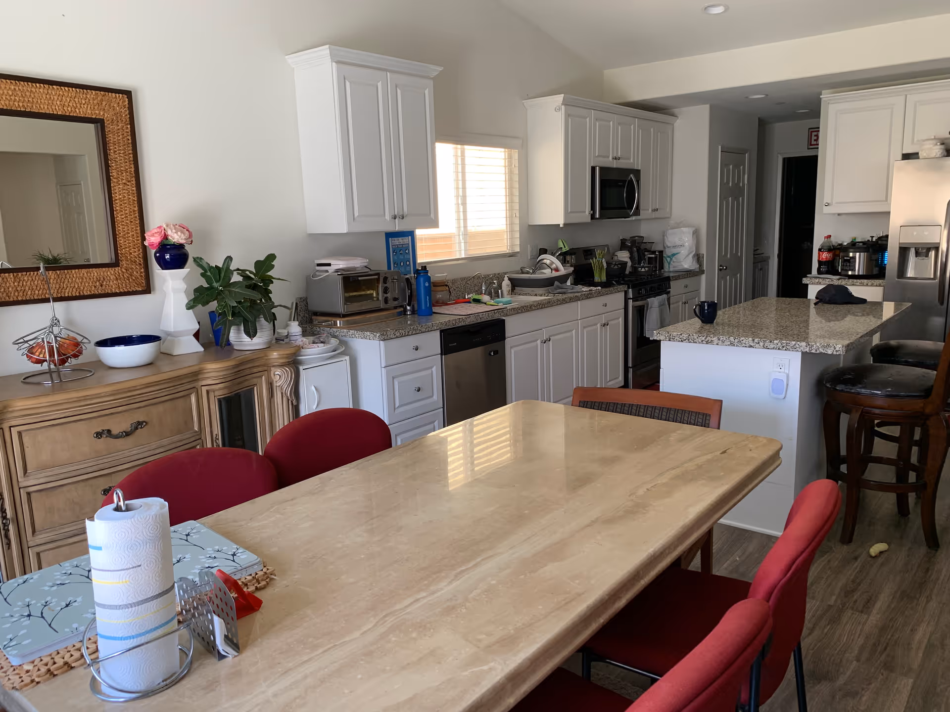 Interior view of a kitchen and dining area with a beige marble dining table surrounded by red chairs. The kitchen features white cabinets, a granite countertop island with two wooden bar stools, a stainless steel refrigerator, microwave, and dishwasher. A wooden sideboard with a large mirror, plants, and decorative items is visible on the left side.