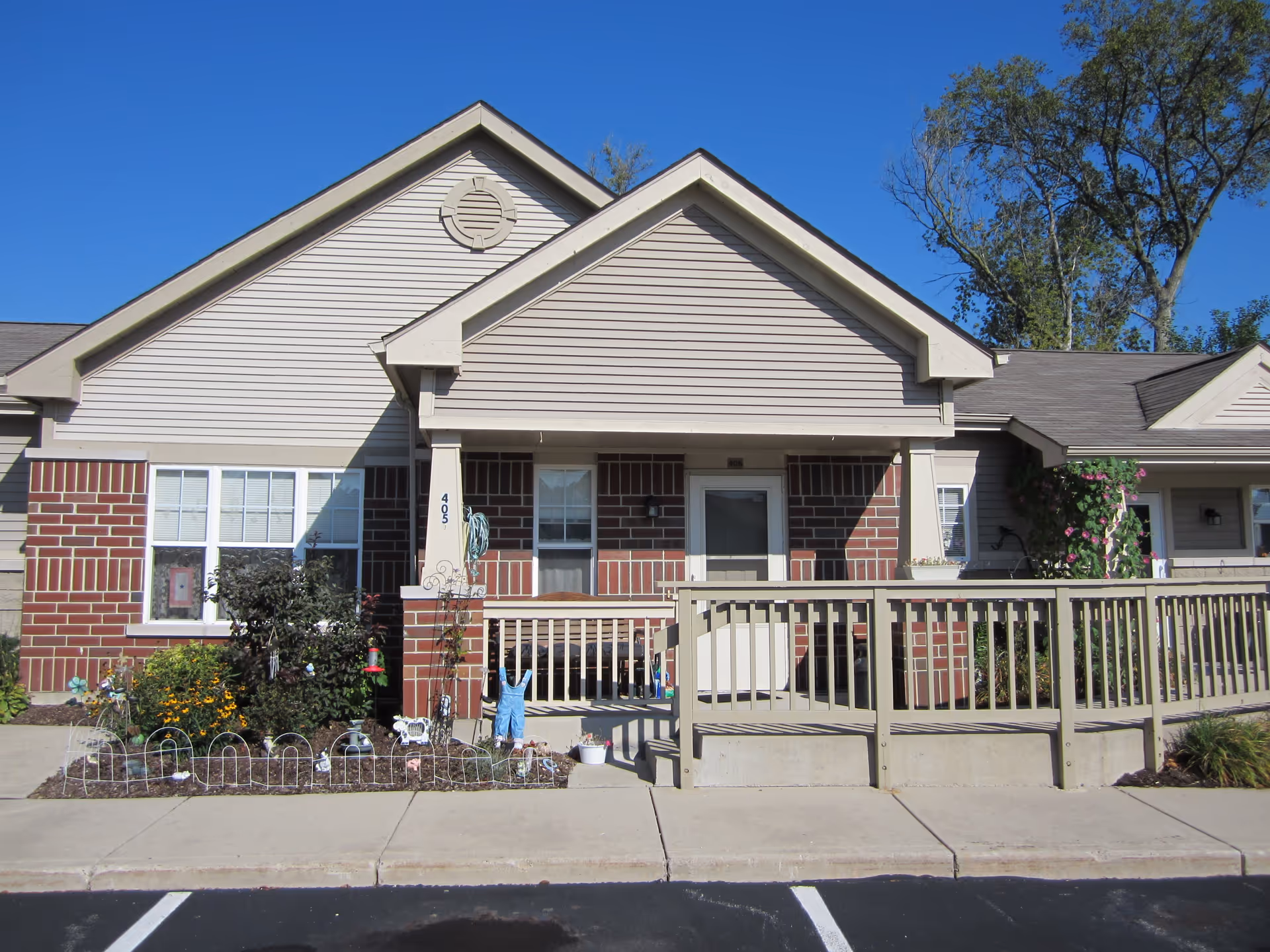 Front exterior of a single-story brick and siding senior housing unit with a covered porch, ramp and small garden.