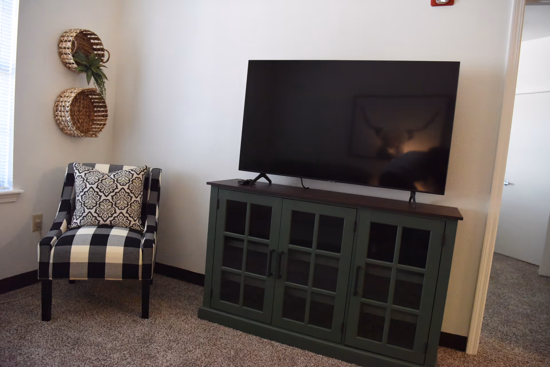 A cozy corner of a living room featuring a black and white checkered armchair with a patterned cushion, two woven wall baskets with a small plant, and a green cabinet with glass-paneled doors supporting a large flat-screen TV. The room has beige carpet and white walls.