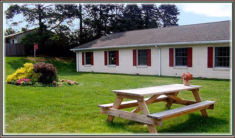 Outdoor area of Cornerstone Living facility showing a wooden picnic table with a flower pot on top, green grass, a flower bed, and a single-story building with white walls and red window shutters in the background.