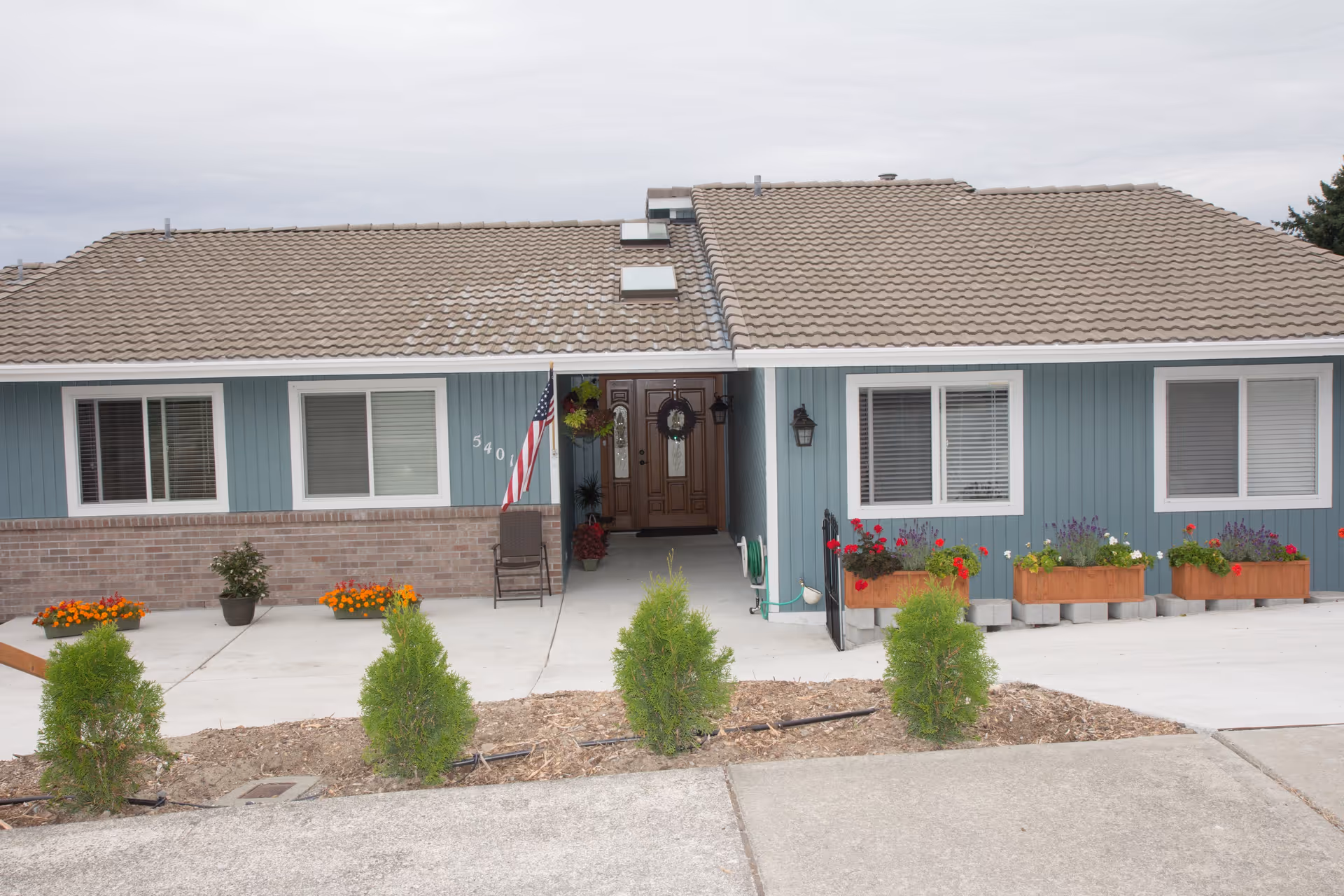 Single-story blue-sided house front with a tiled roof, an American flag by the entrance, potted flowers and window planters.