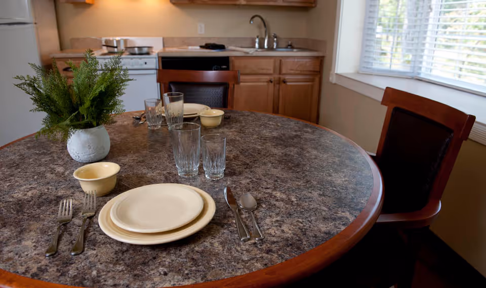 Round dining table set with plates, utensils, glasses and a potted plant in a small kitchen/dining area with cabinets and a window.