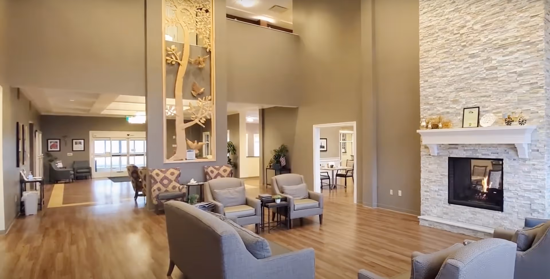 A spacious and well-lit senior living facility common area with wooden flooring, beige walls, and a tall stone fireplace with a mantle decorated with small items. The room features comfortable seating including armchairs and sofas arranged around the fireplace. There is a decorative wall panel with a tree and birds design, and an open doorway leading to another room with a table and chairs. The entrance with glass doors is visible in the background.