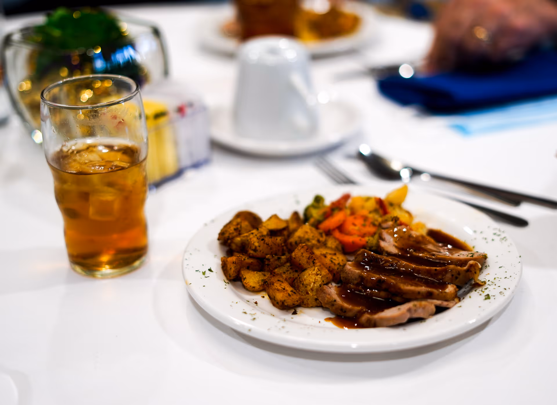 A white plate with sliced meat covered in brown gravy, roasted potatoes, and mixed vegetables on a white tablecloth. Next to the plate is a glass of iced tea with ice cubes. In the background, there are blurred items including a white cup and a blue napkin.