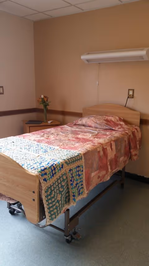 A single hospital-style bed with a wooden headboard and footboard, covered with a patterned quilt and a crocheted blanket at the foot. Next to the bed is a small wooden nightstand with a vase of flowers and a book. The room has beige walls and a linoleum floor.