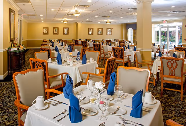 A spacious dining room with multiple tables covered in white tablecloths, each set with blue folded napkins, glassware, plates, and silverware. The room features wooden chairs with cushioned seats and backs, patterned carpet, framed artwork on the walls, and large windows letting in natural light.
