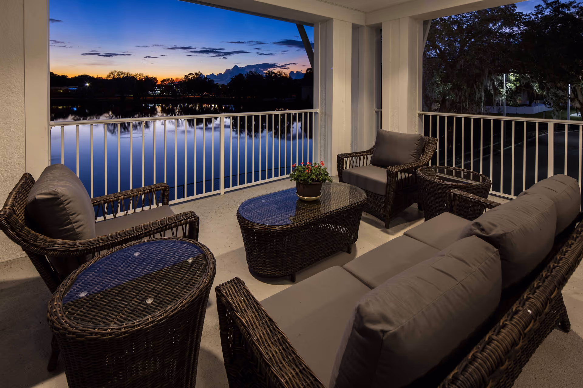 A covered outdoor patio area at dusk with wicker furniture including a sofa, two armchairs, and two glass-topped tables. A small potted plant with red flowers is on the central table. The patio overlooks a calm body of water with trees and a colorful sunset sky in the background.