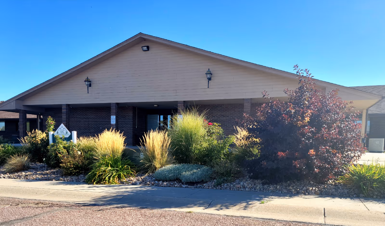 Front entrance of a single-story assisted living building with a gabled roof and landscaped shrubs and ornamental grasses.