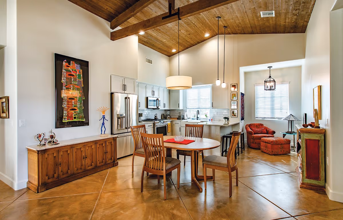 Interior view of a senior living facility showing a dining area with a round wooden table and four chairs, a kitchen with white cabinets and stainless steel appliances, and a cozy sitting area with a red armchair and matching ottoman. The room has a high wooden ceiling with exposed beams and pendant lighting, polished concrete floors, and decorative artwork on the walls.
