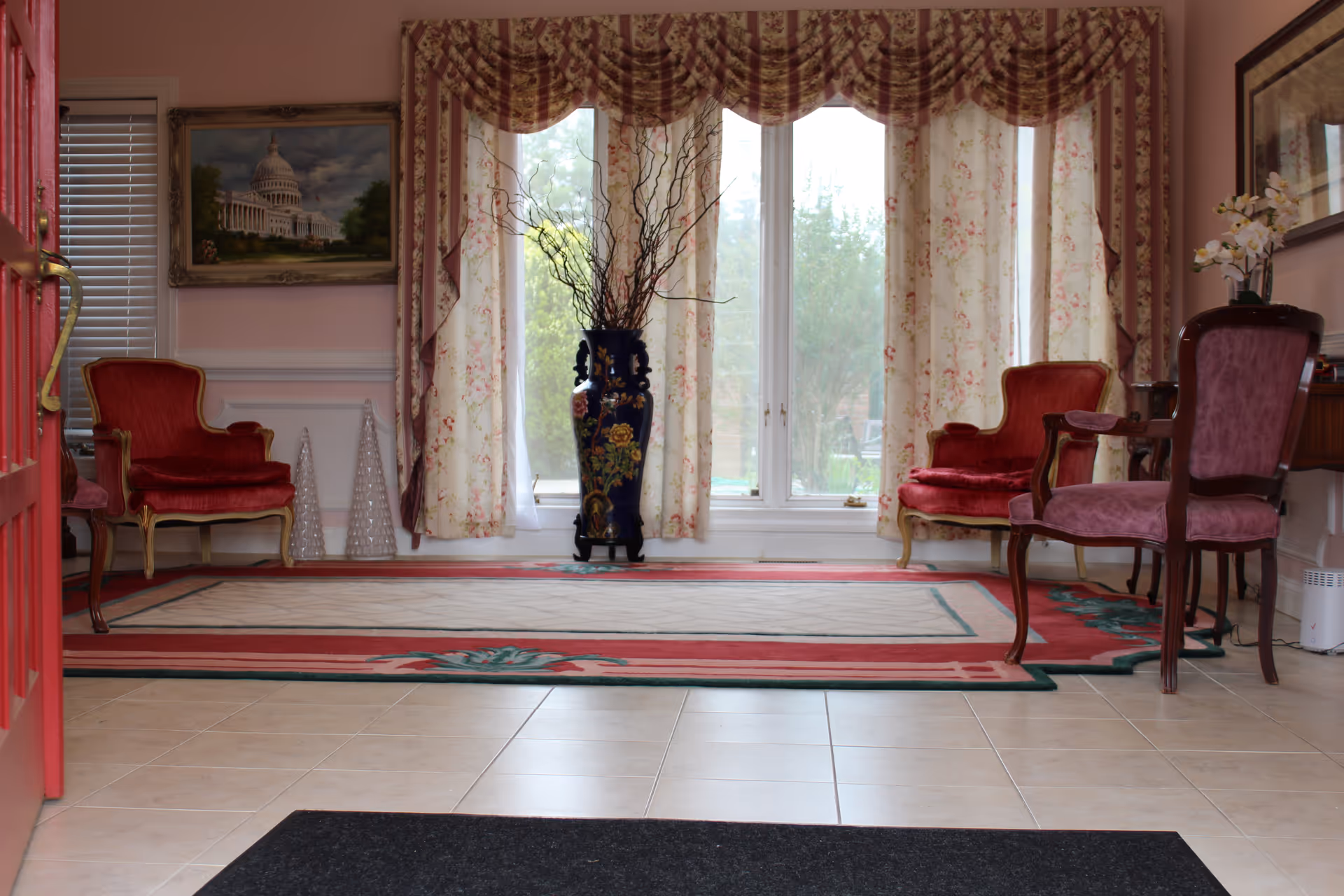 A cozy sitting area with two red upholstered armchairs and one purple upholstered armchair arranged around a large floral-patterned rug. Behind the chairs are large windows with floral curtains and a tall decorative vase with branches in the center. The walls are painted light pink and adorned with framed artwork. The floor is tiled, and a red door is partially visible on the left side.