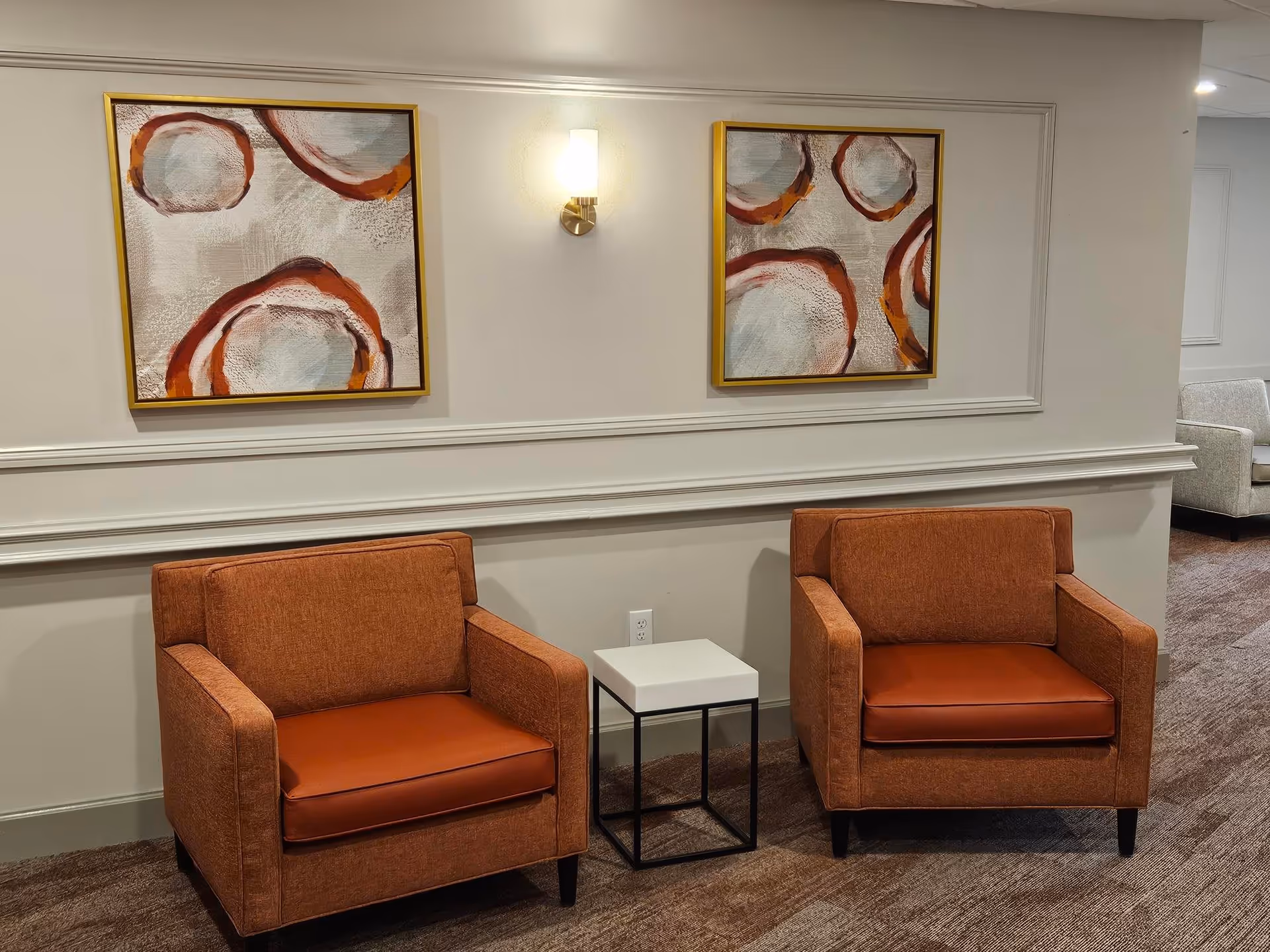 Two orange armchairs with leather seat cushions are placed side by side with a small white square table between them. Above the chairs, two abstract paintings with circular patterns in shades of orange, red, and gray hang on a light-colored wall with decorative molding. The floor is carpeted in a brown tone, and a wall sconce light is mounted between the paintings.